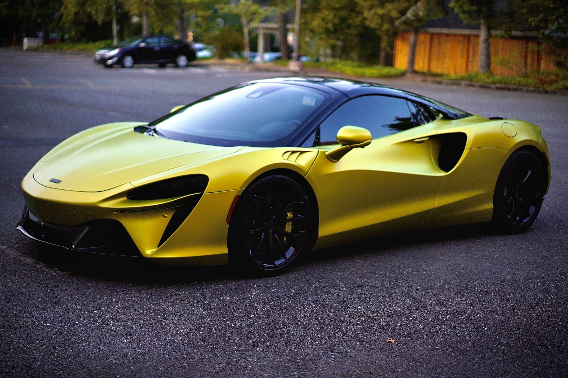 A satin yellow McLaren Artura parked on dark asphalt among trees.